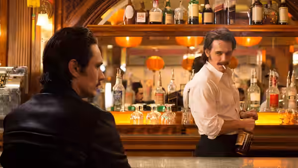Two men at a bar one in leather jacket the other behind the counter holding a bottle
