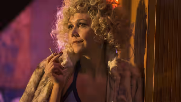 Woman with curly blonde hair in a fur coat holding a cigarette and looking upwards