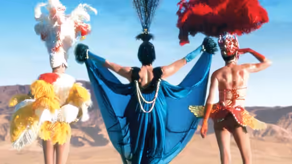 Three drag performers in feathered costumes posing with arms out in the desert