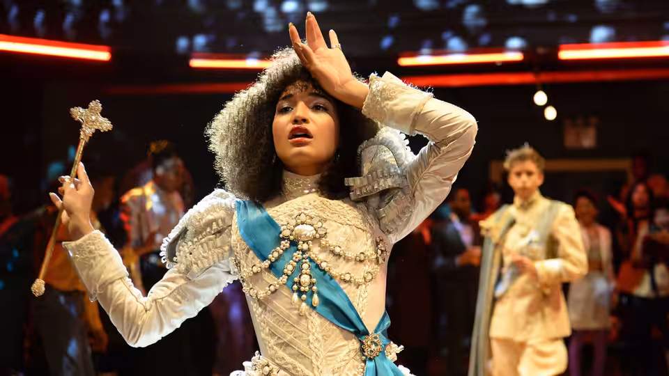 performer in white gown and fur hood raising hand while holding scepter on stage