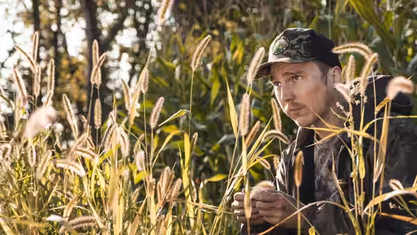 David Sweat (Paul Dano) crouching in tall grass examining something in his hands