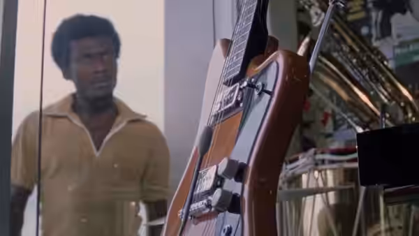 A man in a brown shirt looks toward a guitar displayed in a shop