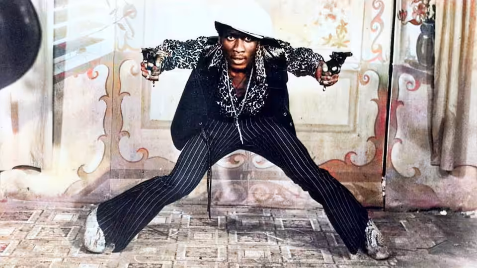 Ivan Martin (Jimmy Cliff) spreads his legs and aims a pistol inside a decorated room