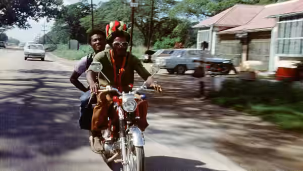 Ivan Martin (Jimmy Cliff) rides a motorcycle with a passenger along a suburban road