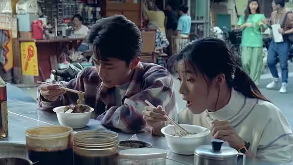 A young man and woman eat noodles at a crowded street-side food stall