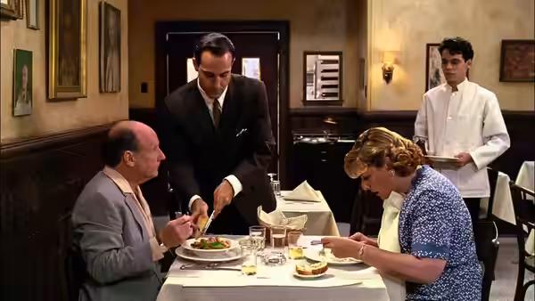 Pascal (Ian Holm) leaning over a table to serve a seated couple in the restaurant