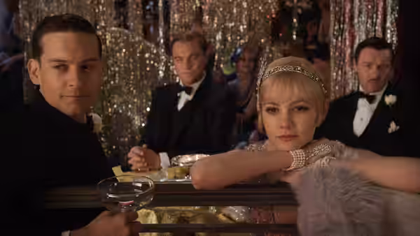Tom Buchanan and Daisy Buchanan lean at a glittering party table as Gatsby watches behind them
