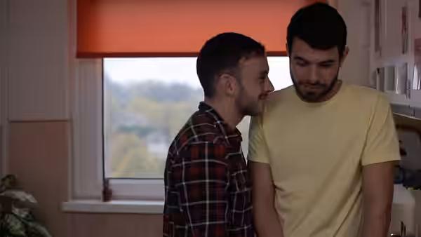Two men standing close together indoors by a window
