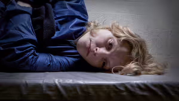 A blonde woman in blue clothing lies on her side on a narrow bed