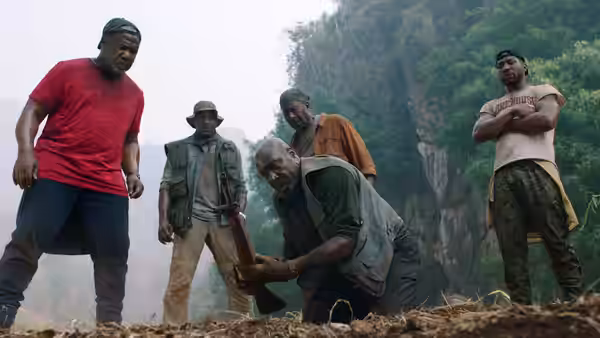 Paul kneels in the dirt holding a wooden object as four men stand around him
