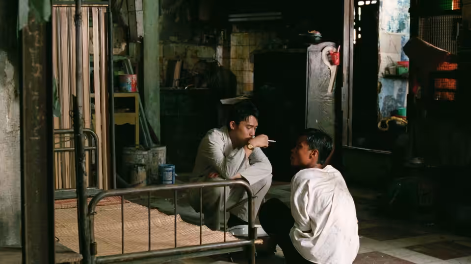 A crouching man speaks to another man seated beside a metal bed in a cramped room