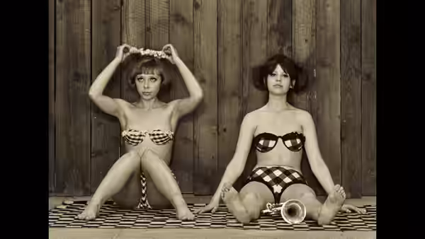 Two women in patterned swimsuits sit on a tiled floor against a wooden wall