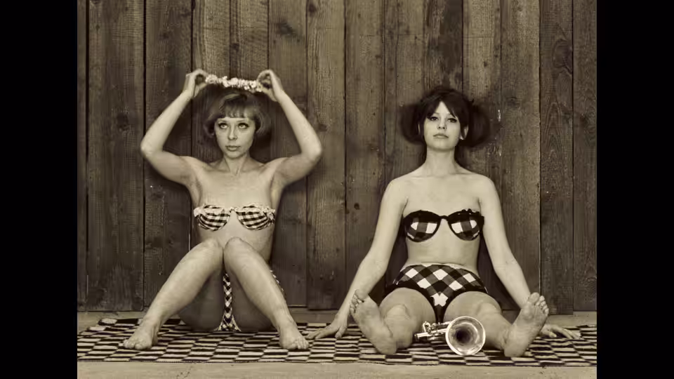 Two women in patterned swimsuits sit on a tiled floor against a wooden wall
