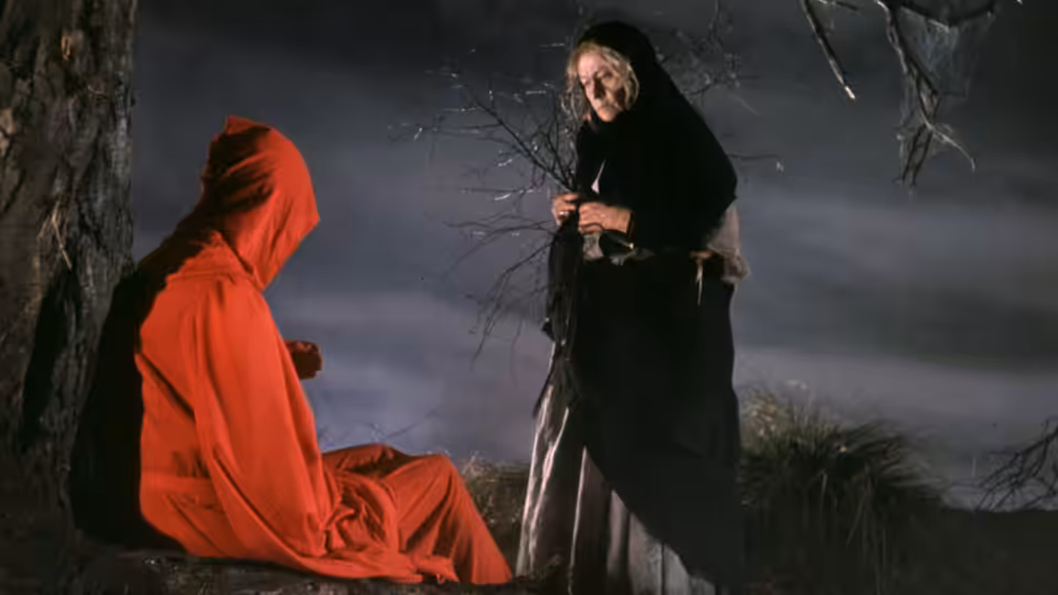 A red-hooded figure sits beside a black-cloaked man beneath bare branches