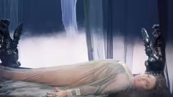 A woman in a pale gown lies on a bed between dark statues