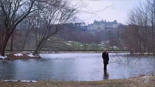 Chance (Peter Sellers) standing in shallow water with a large mansion on the hill behind
