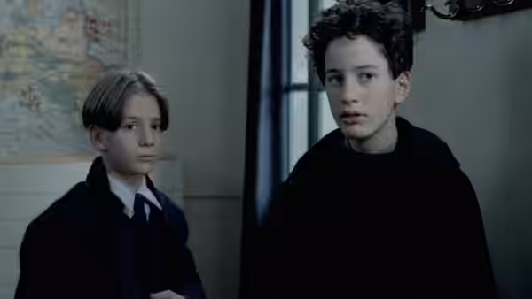 Two boys stand indoors one wearing a black robe near a window