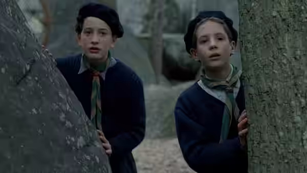 Two boys in berets and scout uniforms peer from behind trees and rocks