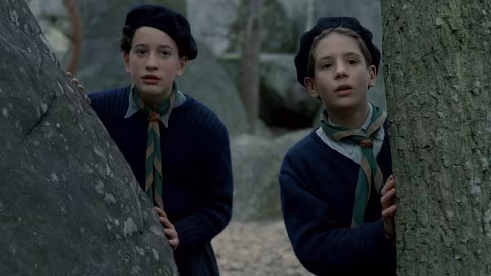 Two boys in berets and scout uniforms peer from behind trees and rocks