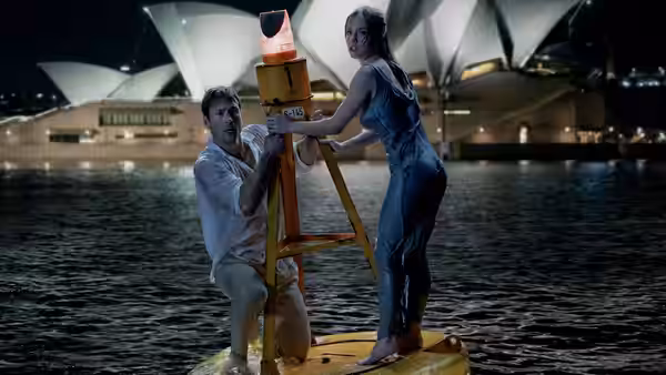 Bea (Sydney Sweeney) and Ben (Glen Powell) on a raft holding a light near the Opera House at night