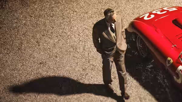 Enzo Ferrari (Adam Driver) standing beside a red racing car on gravel
