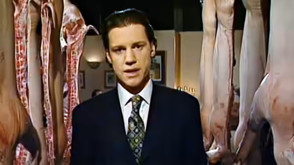 A man in a suit stands among hanging carcasses of meat