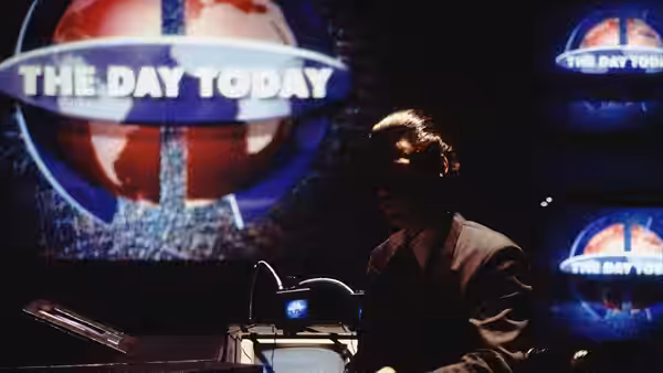 A news anchor works at his desk in front of The Day Today logo