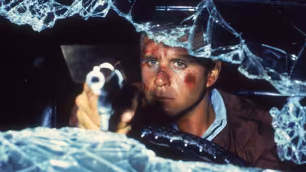 Tom Walsh (Gary Swanson), bruised and bloodied, aims a gun through a shattered windshield