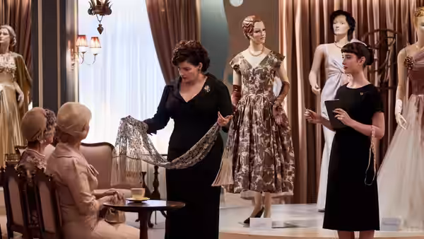 A woman displays a scarf to seated customers as models stand among dresses