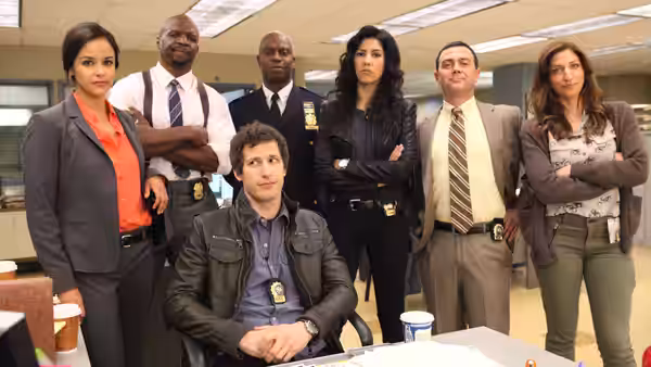 Detectives stand in a police precinct office around a seated colleague