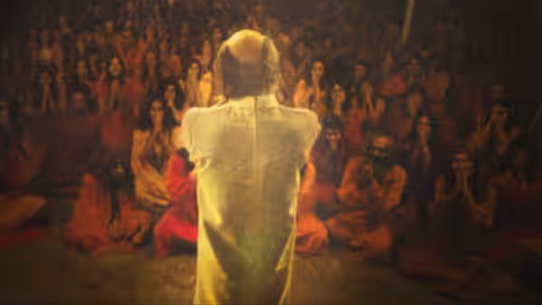 A man in white robes faces a vast crowd of orange-clad followers with raised hands