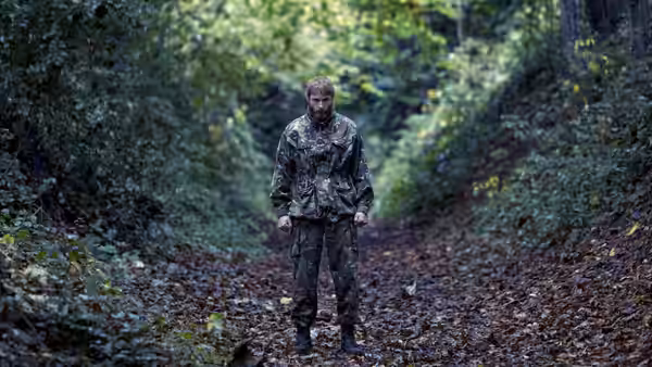 Man in full camouflage stands on a leaf-covered forest path