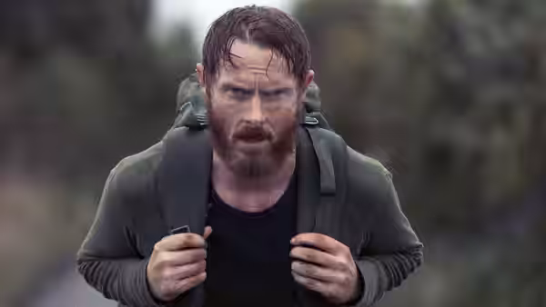 Bearded man grips backpack straps and stares forward with intense expression