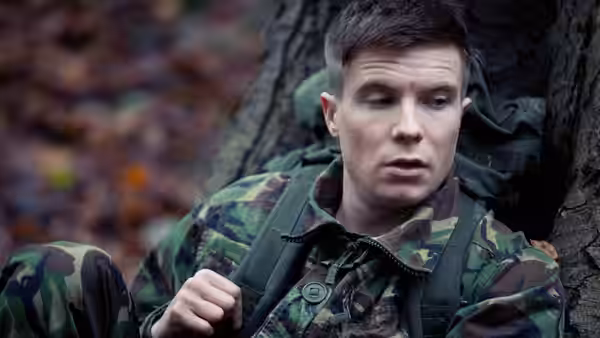 Young man in camouflage rests against a tree in the woods