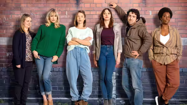 Six people standing in a row against a red brick wall