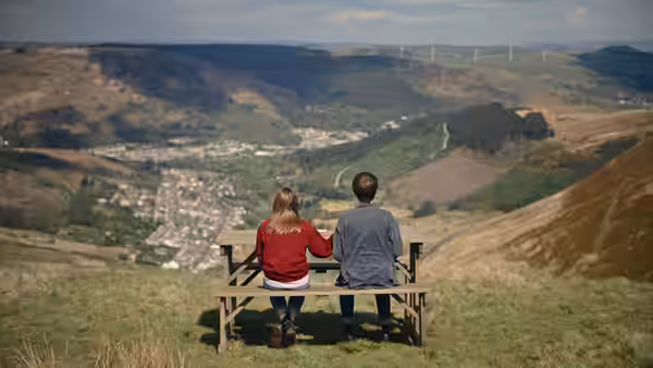 A young couple sits on a bench holding hands overlooking a valley and town