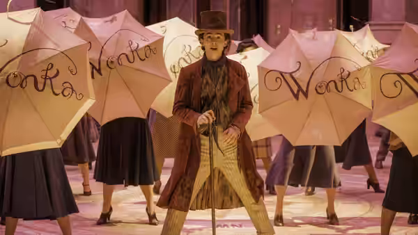 Willy Wonka (Timothée Chalamet) standing among dancers holding pink Wonka umbrellas