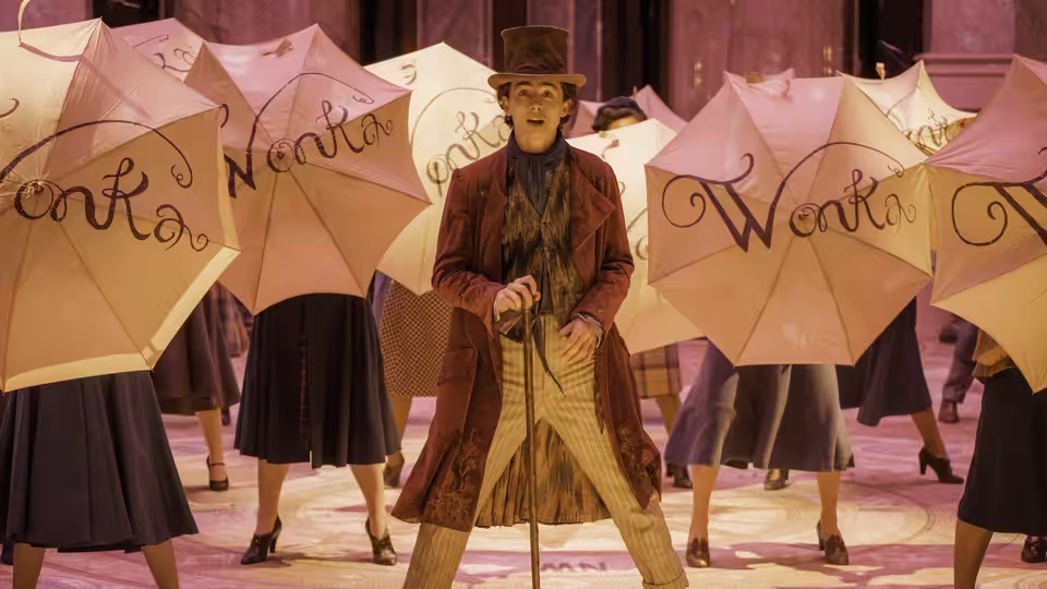 Willy Wonka (Timothée Chalamet) standing among dancers holding pink Wonka umbrellas