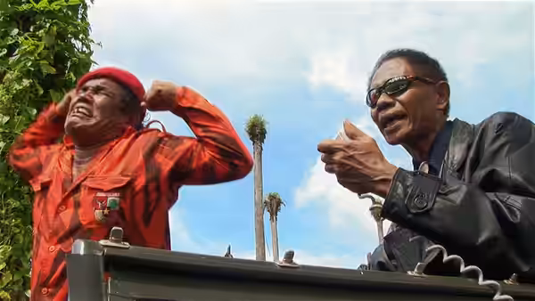 Two men stand on a platform, one gesturing while wearing a red beret and jacket
