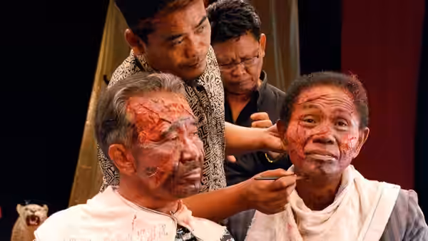 Men apply theatrical makeup to create bloody facial wounds on an older performer