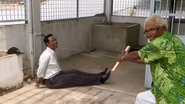 Two men hold wooden objects connected by string while seated in a concrete yard