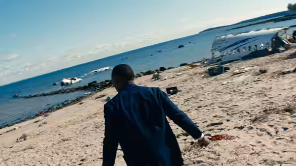 Man in blue suit walking on beach toward crashed airplane wreckage