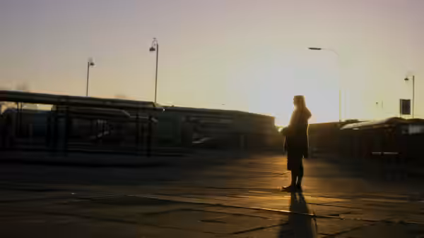 A woman stands alone at a bus terminal as the sun sets