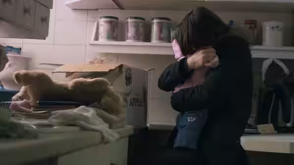 A woman embraces a child beside a counter piled with toys and household goods