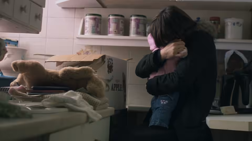 A woman embraces a child beside a counter piled with toys and household goods