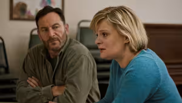 Jay (Jason Isaacs) watches Gail (Martha Plimpton) speak in a room lined with chairs