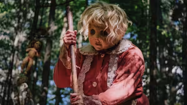Dolly in a red dress grips a wooden bat in a forest with hanging dolls