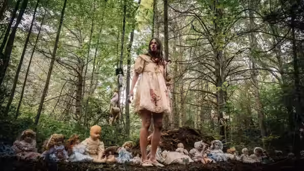 A woman in a bloodied dress stands among many dolls in a forest