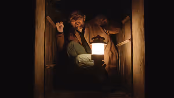 Ohm Bauman (Adam Scott) climbing through a narrow wooden passage with a lantern