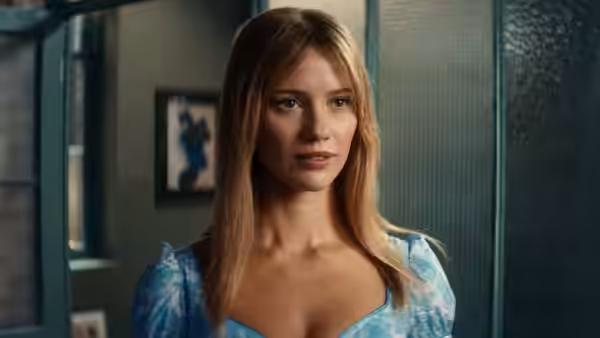 A blonde woman in a blue dress gazes forward with a neutral expression